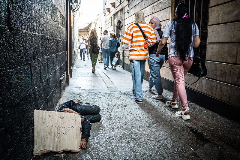 Das neue Syrien: Damaskus. eine Gasse in der Altstadt. Es wird geschätzt, dass noch rund neunzig Prozent der syrischen Bevölkerung in Armut lebt. Und diese Armut ist überall sichtbar, man sieht viele Bettler, darunter auch viele Kinde. Hier: Ein Junge schläft auf dem Boden, bettelt. Passanten gehen meist achtlos an ihm vorbei. Nur wenige geben Geld. Auf dem Schild steht: "Im Namen Gottes, bitte helft mir. Ich möchte mir Kleidung für das Fest kaufen". Das Foto entstand kurz vor dem islamischen Opferfest.